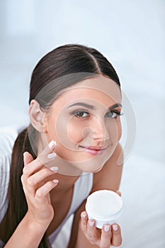 Woman Applying Facial Cream On Face Skin In White Interior