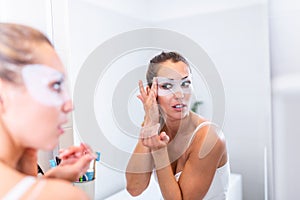 Woman applying mask moisturizing skin mask on face looking in bathroom mirror. Girl taking care of her complexion layering