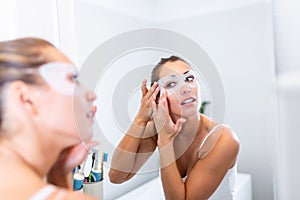 Woman is applying sheet mask on her face in the bathroom. Skin care girl touch patches of fabric mask under eyes to reduce eye