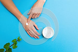 Woman applying white moisturizer on her hands with blue background.