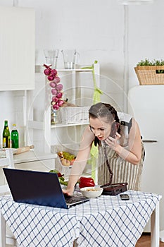 Woman with notebook and phone cooks in kitchen. Isolation period, quarantine, social distancing. Remote education or remote work