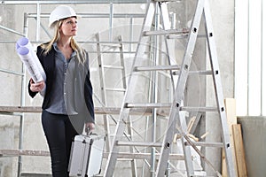 Woman architect or construction engineer bring under arm blueprint wear helmet inside a building site scaffolding and ladder in