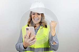 Woman architect or engineer wearing hardhat and reflecting jacket, holding smatphone and making fist in victory.