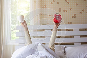 Woman arm hands holding coffee cup and blue alarm clock in on bed in bed room. Young girl with two hands holding blue clock and
