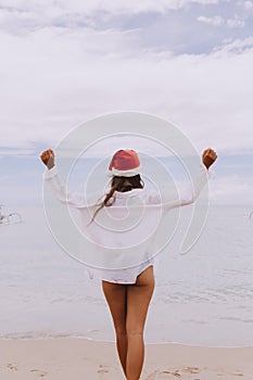 Woman with arms outstretched jumping at beach. Rear view of female is wearing Santa hat and bikini.