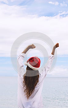 Woman with arms outstretched jumping at beach. Rear view of female is wearing Santa hat and bikini.