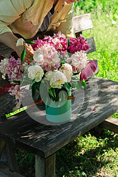 Woman arranging bouquet of peony flowers in milk can on wooden garden bench