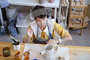 Woman artisian in pottery studio drawing on clay handmade teapot, female ceramist at work
