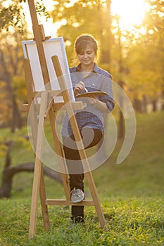 Woman artist drawing a picture on an easel in nature, a girl with a brush and a palette, a concept of creativity and a hobby