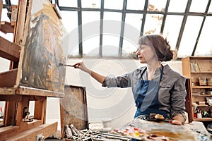 Woman artist paints a picture on canvas. Bright art studio with a large window. Easels and canvases.