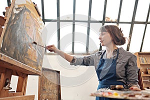 Woman artist paints a picture on canvas. Bright art studio with a large window. Easels and canvases.