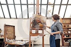 Woman artist paints a picture on canvas. Bright art studio with a large window. Easels and canvases.