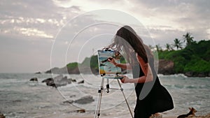 Woman artist paints seascape on canvas at dusk on stormy beach. Waves crash as creator works with palette, brush