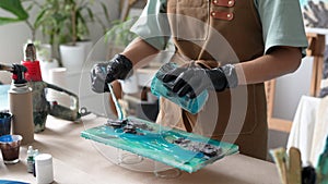 Woman artist pours epoxy resin and smears it with stick on wooden board, using liquid art technique