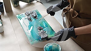 Woman artist pours epoxy resin and smears it with stick on wooden board, using liquid art technique.