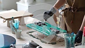 Woman artist pours epoxy resin and smears it with stick on wooden board, using liquid art technique