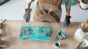 Woman artist pours epoxy resin and smears it with stick on wooden board, using liquid art technique.