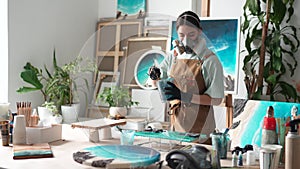 Woman artist in respirator examines painting after pouring epoxy resin, check quality of work