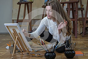 Woman artist sits on the floor among easels with paintings in an art studio and paints an oil painting