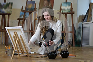 Woman artist sits on the floor among easels with paintings in an art studio and paints on canvas