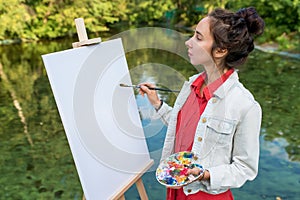 woman artist in summer in park near lake, river pond, draws picture, white canvas, start drawing. Creation of creativity
