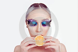 Woman with artistic purple blue eyes makeup feather on eyelashes holding lemon slice in mouth