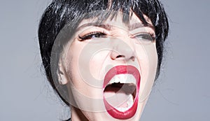 Woman with attractive red lips shouting. Lady in black wig with make up on grey background. Girl on scandalous shouting photo