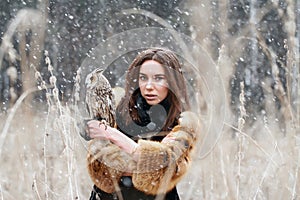 Woman in autumn in fur coat with owl on hand first snow. Beautiful brunette girl with long hair in nature, holding an owl.