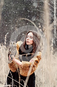 Woman in autumn in fur coat with owl on hand first snow. Beautiful brunette girl with long hair in nature, holding an owl.