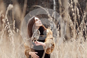 Woman in autumn in fur coat with owl on hand first snow. Beautiful brunette girl with long hair in nature, holding an owl