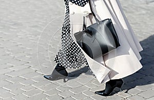 Woman in autumn or spring in a beige coat and a black dress with white polka dots. Fashionable bag close-up in female hands.