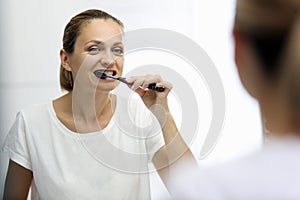 Woman in awhite T-shirt is brushing her teeth in front of mirror