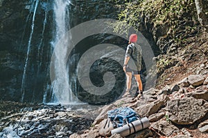 Woman with backpack dressed in red hat and active trekking clothes standing near the mountain river waterfall and enjoying the