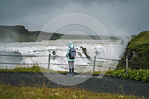 Woman with backpack and green jacket visit Gullfoss powerful famous waterfall in Iceland. Tourist attraction who follow Golden