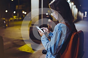 Woman with backpack pointing finger on blank screen smartphone on background bokeh light in night atmospheric city, blogger