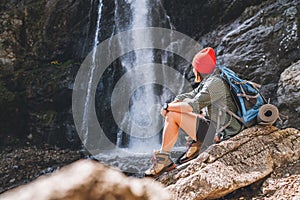 Woman with a backpack in red hat dressed in active trekking clothes sitting near the mountain river waterfall and enjoying the