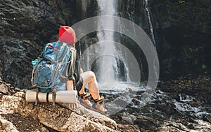 Žena s batohom v červenom klobúku oblečená v aktívnom trekingovom oblečení sedí v blízkosti vodopádu horskej rieky a teší sa