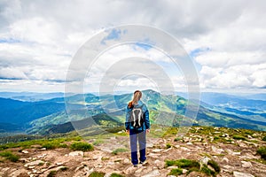 Woman with backpack rise to the mountain top. Girl stands atop a rock just off a hiking trail mountain range back