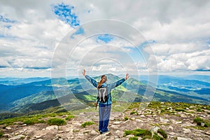 Woman with backpack rise to the mountain top, raises his hands up. Girl atop a rock just off a hiking trail mountain