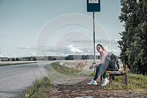 A woman with a backpack sits at a rural bus stop. The tourist is waiting for the bus