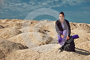 Woman with backpack sitting on one knee on yellow dunes looks into distance on sunny day