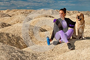 Woman with backpack sitting on yellow dunes with dog labrador retriever looks into distance on sunny day