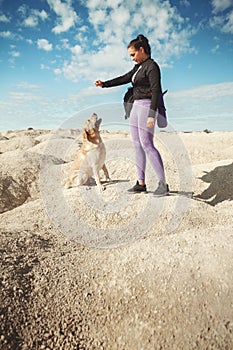 woman with backpack standing on yellow dunes and commands dog labrador retriever and hold him by paw on sunny day