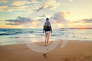 Woman with backpack walk on the ocean sand beach at sunset time