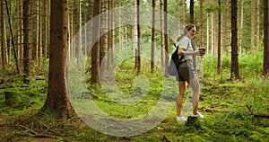 A woman with a backpack walking in a dense pine forest, holding a a pot with a small pine tree seedling for planting it