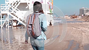 Woman with backpack walks near coast guard post. Hair blowing in wind. Girl walking along ocean beach. Slow motion.