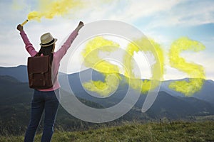 Woman with backpack and word SOS made of color smoke bomb in mountains, back view