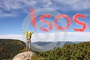 Woman with backpack and word SOS made of color smoke bomb on rocky peak in mountains