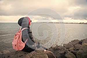 Woman backpacker sit on stone sea coast in reykjavik, iceland. Woman enjoy sea view on cloudy sky. Girl travel with