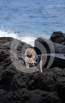 Woman with blonde hair sitting on the rocks near the sea typing her smartphone